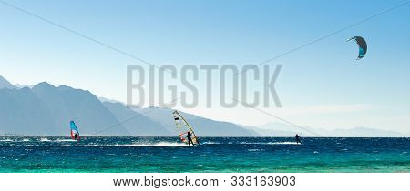 Windsurfers And Kitesurfers Ride In The Red Sea On The Background Of The Rocky Coast In Egypt Dahab