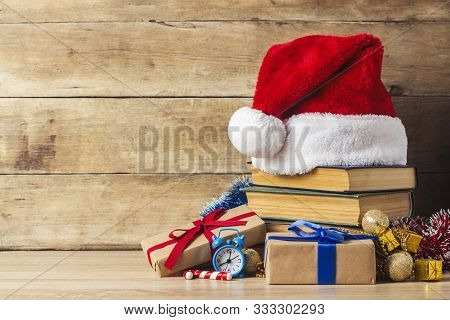 A Stack Of Books, Santa Clauss Cap, An Alarm Clock, Gift Boxes On A Wooden Background. Concept Of Ne