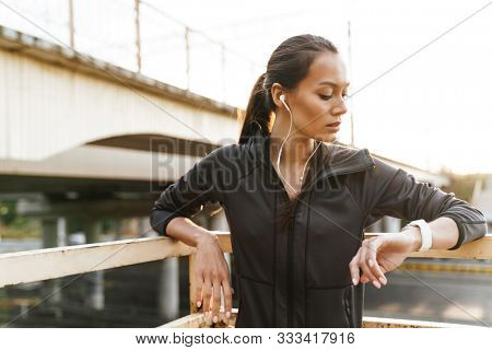 Image of young focused woman using earphones and smartwatch while leaning on railing near road bridge in morning