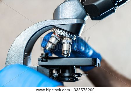 Male Laboratory Assistant Examining Biomaterial Samples In A Microscope. Cllose Up Hands In Blue Rub