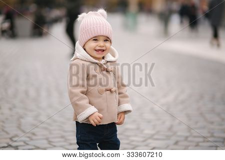 Little Smiled Baby Girl Dressed In Brown Coat Denim Jeand And Pink Hat Stand In Centre On The City. 