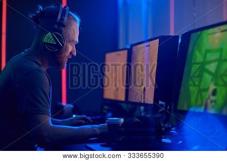 Young Man In Gaming Headphones Looking Seriously He Sitting In Front Of Computer Monitors And Playin