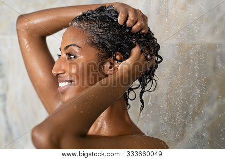 Woman washing hair showering in bathroom at home. Smiling black woman bathing while looking away. Happy woman rinsing hair while taking a shower at luxury spa.