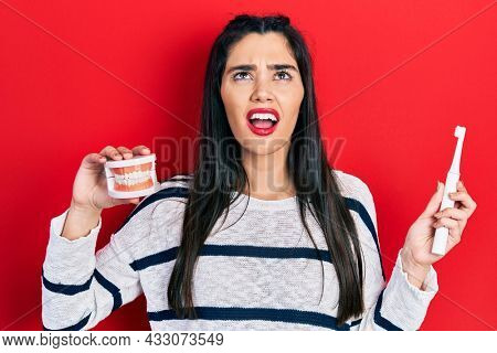 Young hispanic girl holding electric toothbrush and denture angry and mad screaming frustrated and furious, shouting with anger looking up. 