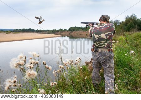 A Hunter Shoots A Flying Duck With A Shotgun. He Stands On The Bank Of The River, In Summer.