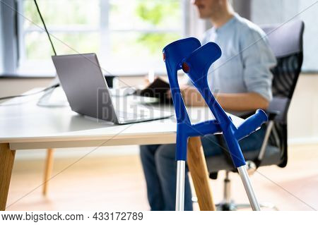 Handicapped Injured Employee With Crutches At Workplace Desk