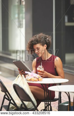 Prety Positive Young Woman Reading News Article On Tablet Computer And Enjoying Refreshing Drink At 