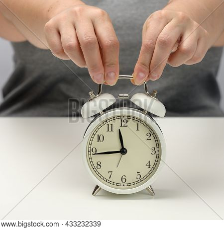 Female Hand Holds A White Metal Alarm Clock On A White Table, The Time Is Fifteen To Twelve. Concept