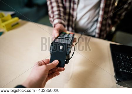 Close-up Shopper Hand Gives Credit Card To Pay Through Payment Terminal, Seller Behind Counter Makes