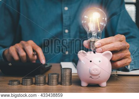 Businessman Hand Holding Lightbulb On Piggy Bank With Money Stack Step Up On Office Desk, Saving Ene