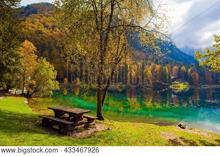 The wooden table with benches. Lake Fuzine. Yellow and orange trees are reflected in the green smooth water of the lake. Alps, Northern Italy 