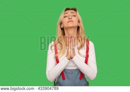 Please, God. Portrait Of Upset Pleading Adult Woman In Denim Overalls Praying With Closed Eyes, Impl
