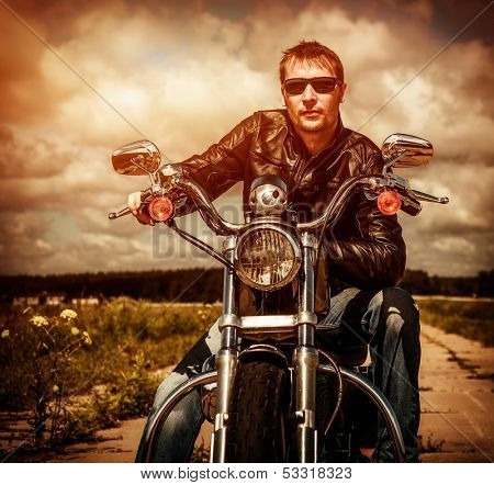 Biker man wearing a leather jacket and sunglasses sitting on his motorcycle looking at the sunset.