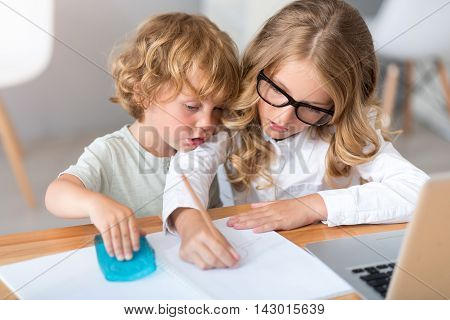 Time together. Serious little girl with glasses drawing in the notebook while the little charming boy sitting near her in front of the laptop