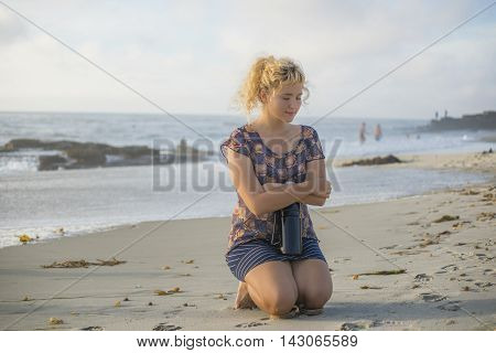 Cold shivering Young blonde woman sitting holding thermos on beach in California with eyes closed