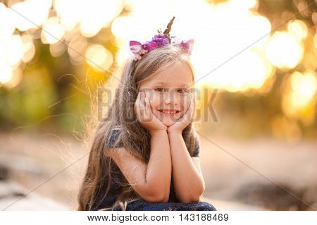 Smiling kid girl 4-5 year old wearing unicorn headband over nature background. Looking at camera. Childhood. Summer portrait over sunset. Selective focus.