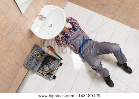 High Angle View Of Male Plumber Lying On Floor Repairing Sink In Bathroom
