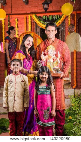 Smart Indian couple holding Ganesh idol or ganpati idol at entrance, welcoming lord ganesh on ganesh festival or chaturthi with kids and parents, happiness concept