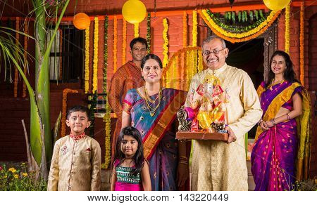 Smart Indian old couple holding Ganesh idol or ganpati idol at entrance, welcoming lord ganesh on ganesh festival or chaturthi with grand childrens and kids in the background, happiness concept
