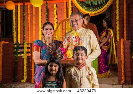 Smart Indian old couple holding Ganesh idol or ganpati idol at entrance, welcoming lord ganesh on ganesh festival or chaturthi with grand childrens and kids in the background, happiness concept