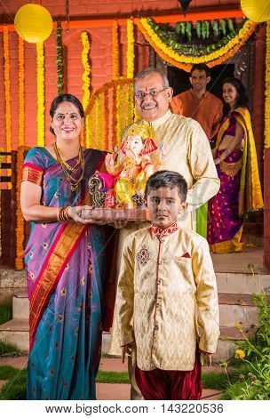 Smart Indian old couple holding Ganesh idol or ganpati idol at entrance, welcoming lord ganesh on ganesh festival or chaturthi with grand childrens and kids in the background, happiness concept