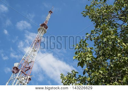 Broadcast transmitting antenna for radio and television signal on green tree and sky background.