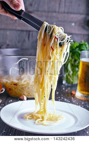 Pasta with gralic and chili. Hand with tongs move spaghetti with garlic, oil and chili to a white plate on wood background