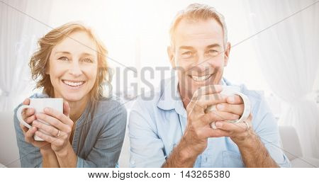 Smiling middle aged couple sitting on the couch having coffee looking at camera at home in the living room