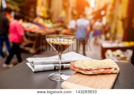 Shakerato drink in cocktail glass with panini and newspaper on the table. Italian lunch with traditional coffee and sandwich with prosciutto