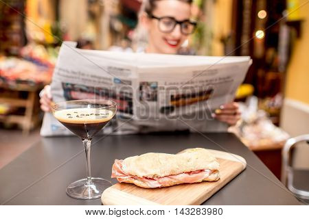 Young woman reading newspaper with shakerato drink and panini on the table outdoors on the street in italian city. Traditional italian lunch. Image with small depth of field, focused on the food