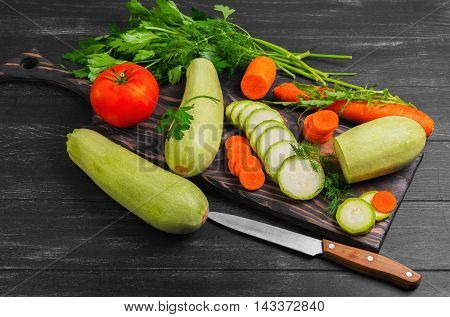 Vegetables ingredients for vegetable dishes. Vegetables marrows white squashes carrots parsley lettuce dill tomato. Marrows white squashes carrots whole and cut into pieces. The dark black wood background.