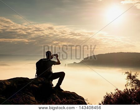Dreamy fogy landscape. Amateur photographer takes impressive photos with phone. Hiker with backpack on cliff photogrphy orange pink misty sunrise in a beautiful valley below