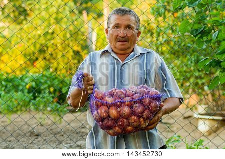 An old man is holding a sack of onions in his hands. He is surprised. He has a good harvest