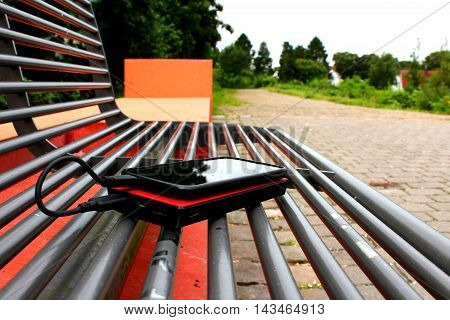 A black smartphone and a red powerbank on a bench in a park