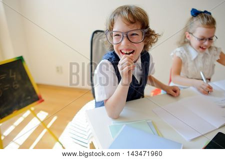 Ridiculous little students bespectacled boy and girl share the same desk. They are pupils of an elementary school. They laugh. On the desk in children are school supplies and textbooks. Back to school