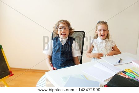 Ridiculous little students bespectacled boy and girl share the same desk. They are pupils of an elementary school. They laugh. On the desk in children are school supplies and textbooks. Back to school