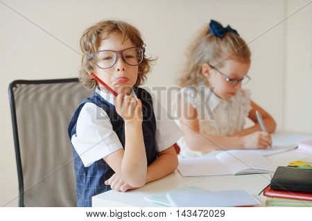 Funny little pupils sit at one desk. They are pupils of an elementary school. Boy fools around girlie writes something with a serious look. On a school desk children have school accessories and textbooks.