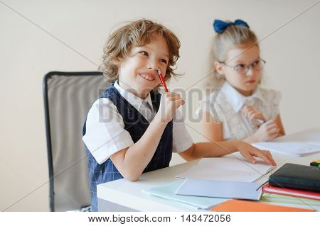 Funny little pupils sit at one desk. They are pupils of an elementary school. Boy fools around at girl a serious look. On a school desk children have school accessories and textbooks. Back to school.