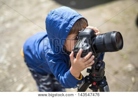Small kid in a blue windcheater is making a photo shoot on the blurry background outdoors. He is looking into the viewfinder of the camera which is on the tripod. Horizontal.