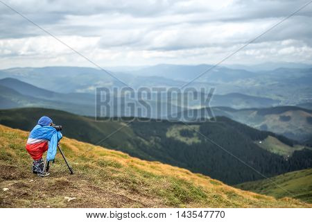 Cute kid in multi-colored big size windcheater is making a photo on the background of mountains and the cloudy sky. He is looking into the viewfinder of the camera which is on the tripod. Horizontal.