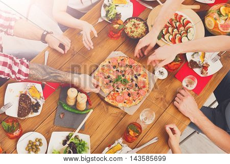 People eat pizza at festive table served for party. Friends celebrate with catering food on wooden table top view. Woman and man's yands take the dish with italian pizza.