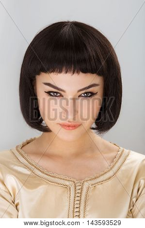 High fashion look. Glamorous closeup portrait of beautiful sexy stylish brunette young woman model with bright makeup with perfect clean skin in gold dress posing against bright gray background. Cleopatra