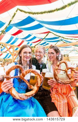 Three friends in beer tent at Dult or Oktoberfest holding giant pretzels up in the air
