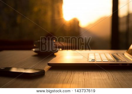 Nomad Work Concept Image Computer Coffee Mug and Telephone on black wood Table and Evening Sunlight shining throw large windows focus on Edge of Laptop