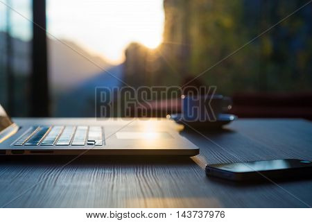 Nomad Work Concept Image Computer Coffee Mug and Telephone on black wood Table and Evening Sunlight shining throw large windows focus on Edge of Laptop