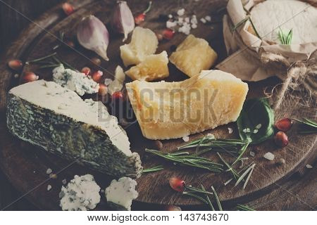 Cheese delikatessen closeup on rustic wood. Wooden desk with blue cheese roquefort, parmesan, camembert and brie cuts decorated with garlic, pomegranate and rosemary. Soft toned candid image