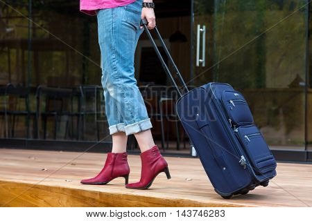 Person walking on wooden terrace pulling Travel Suitcase female Legs and Hand casual vacation Clothing red attractive high Heels Shoes and Jeans