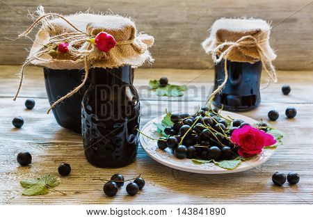 Preserved homemade black currant jam in glass jars on light wooden table. Fresh berries and green leaves white vintage plate.
