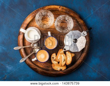 Coffee espresso in cups with italian cantucci, cookies and milk in jug on wooden serving round board over dark blue painted plywood background, top view, horizontal composition
