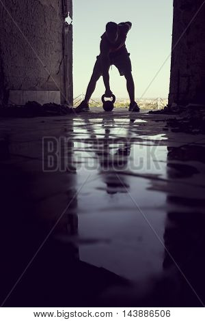 Muscular athletic built young athlete working out in a ruin building next to a puddle of water. Crossfit training with barbells and kettlebell deadlifting